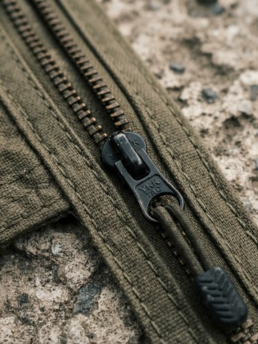 Macro close-up of heavy-duty tactical zippers and olive-drab double-stitched seams on weathered concrete, hard overcast daylight, desaturated earth tones, 35mm