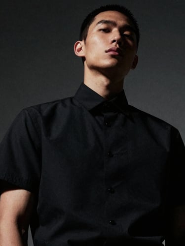 Dramatic low-angle portrait of a model wearing a structured matte black short-sleeve shirt, high-contrast directional lighting, deep shadows, desaturated tones, 35mm film grain