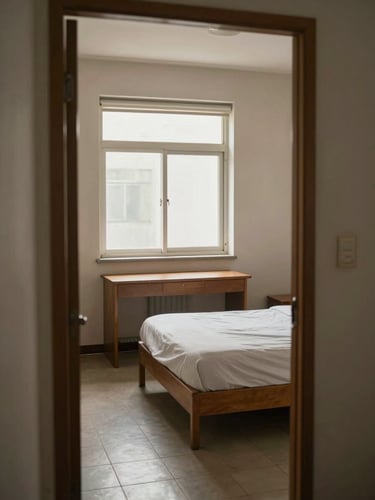 Wide doorway-perspective shot of a furnished PG room interior — single bed with plain sheets, wooden desk beneath a window, tiled floor, neutral walls — natural midday window light, no people, no staging