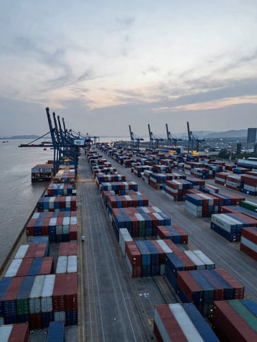 Wide panoramic aerial shot of Shanghai Yangshan deep-water port at dusk, hundreds of shipping containers stacked in precise rows extending to the horizon, cool blue-grey natural light, port cranes silhouetted against a pale overcast sky, shot from high altitude with the container yard filling the right two-thirds of the frame and open water visible at the left edge