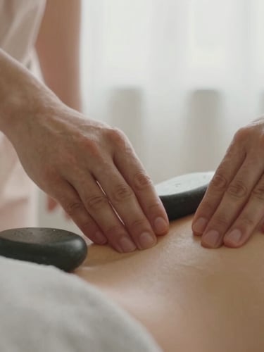 Close-up of professional hands performing a massage with warm stones on a clean, linen-covered back, soft diffused window light, warm sandy tones, shallow depth of field, 35mm