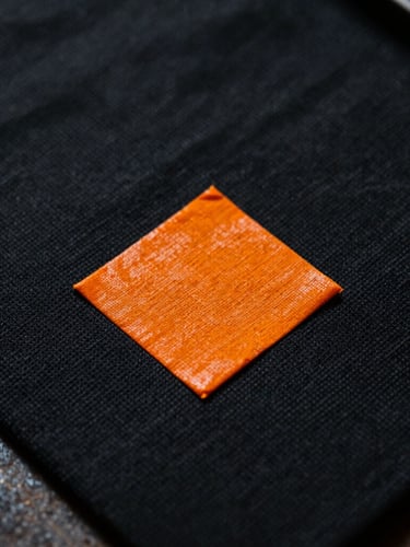 Macro close-up of thick safety-orange ink curing on a black heavyweight cotton canvas, dramatic side-lighting, gritty workshop texture.