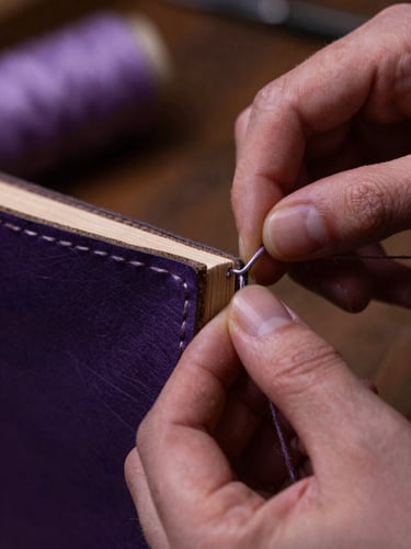 Macro shot of hands sewing a leather book spine with thick linen thread, warm low-key workshop lighting, deep amethyst and lavender tones, tactile textures, 35mm macro lens