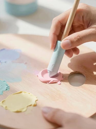 Close-up of hands mixing soft pastel pigments on a wooden palette, warm diffused sunbeams casting gentle shadows, airy whimsical tones.
