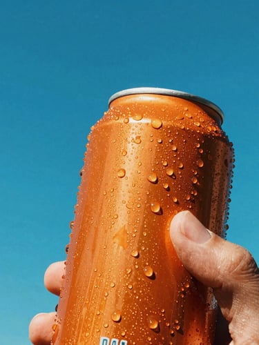 Low-angle heroic close-up of a condensation-dripping orange soda can held by a hand, direct harsh sunlight, sharp shadows, vibrant outdoor background