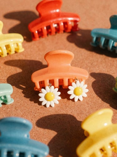 Close-up flat lay of colorful retro hair claws and sun-drenched daisy earrings on a textured terracotta surface, bright natural golden-hour sunlight, 35mm film look.