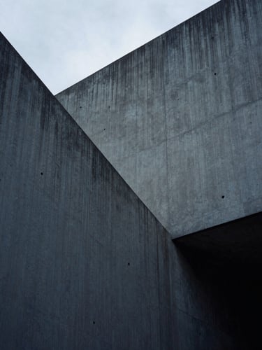 Low-key cool-toned photograph of sharp concrete architectural angles, deep shadows, overcast diffused daylight, 35mm