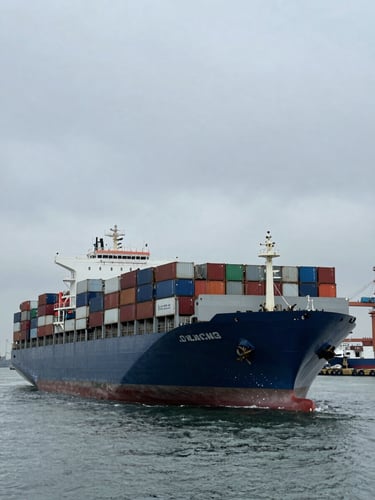 Wide environmental shot of a massive container ship loaded with cargo and vehicles leaving a busy industrial port, dramatic overcast daylight, desaturated steel and blue tones, 35mm