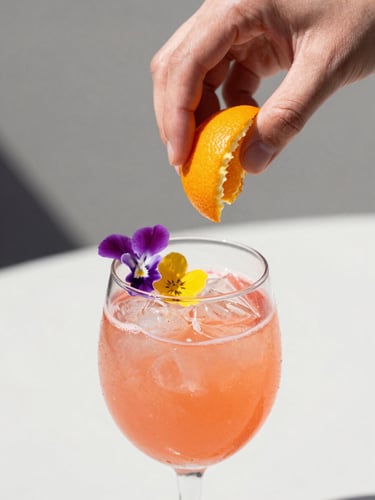 Close-up shot of hands expressing a fresh orange peel over a vibrant pink cocktail garnished with edible wildflowers, bright natural daylight, shallow depth of field