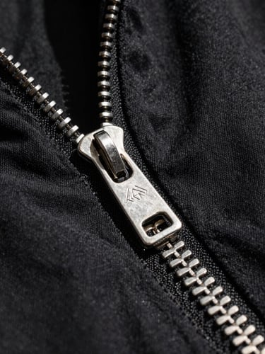 Extreme close-up of a high-contrast metallic silver zipper on a raw black upcycled cotton jacket, direct sunlight casting sharp shadows, macro texture.