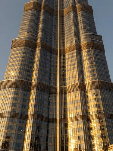 Symmetrical wide-angle architectural shot of a soaring Dubai skyscraper facade during golden hour, intense warm sunlight reflecting off black glass and brushed gold metallic panels, sharp focus, 35mm.