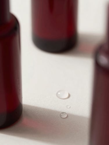 Close-up of elegant product bottles styled alongside clean water droplets and soft shadows, minimalist editorial photography, soft natural side-light, deep burgundy tones, macro lens detail.