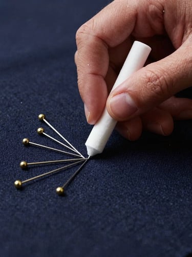 Macro close-up of a tailor's hands marking dark navy wool fabric with white chalk, brass pins neatly arranged nearby, soft side lighting highlighting the textile texture