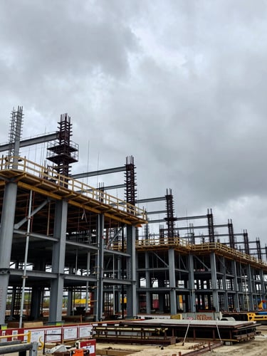 Wide environmental shot of an active industrial construction site with structural steel framing under an overcast gray sky, high contrast.
