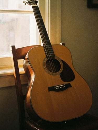 Warm vintage film photography of a worn acoustic guitar leaning against a wooden chair, dusty golden-hour light streaming through a window, 35mm grain.