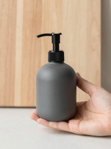 A hand holding a matte charcoal soap dispenser next to a light wood cutting board, soft diffused daylight, warm neutral tones, clean composition, 35mm lens, minimalist bathroom setting