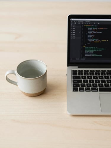 Overhead flat-lay of a minimalist wooden desk in soft morning daylight, a single ceramic teacup next to a sleek laptop displaying clean code syntax highlighting, warm cream tones.