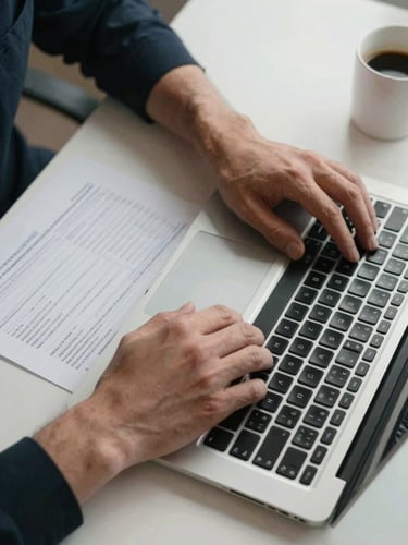 Close-up overhead framing of hands resting on a laptop keyboard on a wide desk, a printed reconciliation sheet visible to the left, overhead office fluorescent light casting clean even shadows, a half-empty coffee mug at the far edge