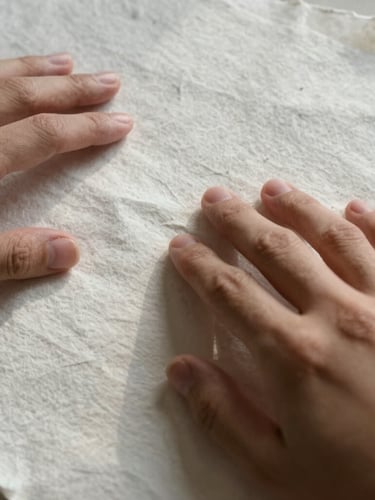 Intimate close-up of hands touching textured heavy cotton paper with soft-edged mountain daylight casting gentle shadows, showing the raw fiber edges.