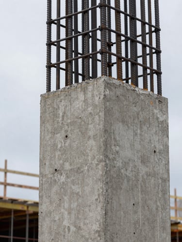 Close-up detail of a reinforced concrete structural column at an active construction site in Mexico — steel rebar emerging from formwork, natural overcast daylight from the left, cool grey tones, no people, tight framing on the material texture and engineering geometry