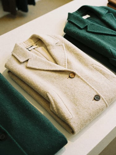 Editorial fashion photography of neatly folded premium wool coats in a warm cream and forest green tone, soft natural gallery lighting, medium format film