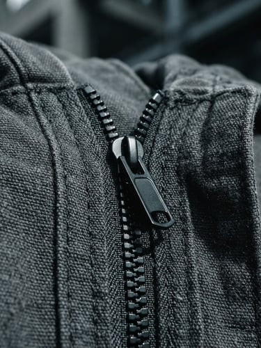 Extreme close-up of heavy charcoal cotton fabric with reinforced stitching, a matte black technical zipper glints under cool overcast daylight, sharp geometric shadows.