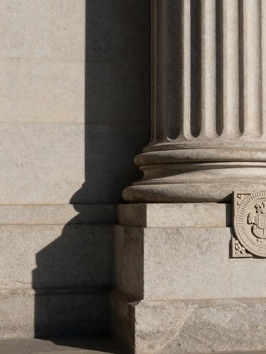 Close-up architectural shot of a courthouse exterior column base, deep shadow and worn stone texture under flat midday light, official carved seal partially visible at the right edge, generous negative space of stone wall to the left