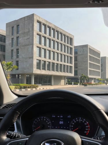 First-person driver-seat perspective of a clean SUV steering wheel, looking out at the modern concrete architecture of a Bangalore tech park in bright morning light