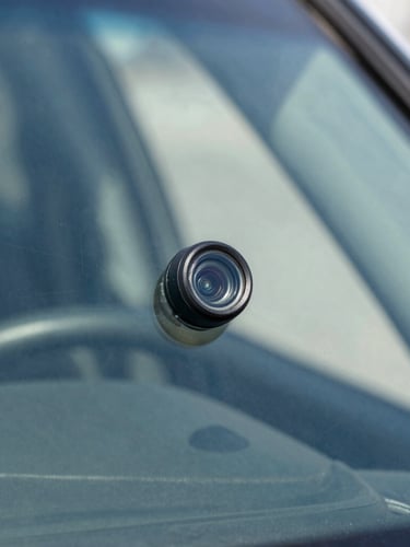 Macro close-up of a high-end vehicle rearview mirror assembly showing the camera sensor lens flush against clean windshield glass, sharp focus, 35mm
