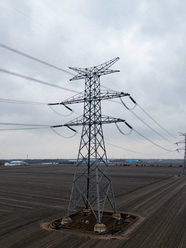 Low-angle heroic shot of high-voltage transmission towers cutting across a vast, flat, graded industrial parcel under a high-contrast overcast sky