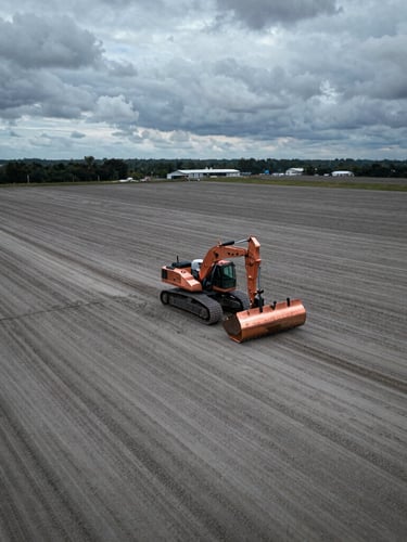 Aerial drone shot of a massive flat graded land parcel with heavy construction grading equipment visible, sharp copper accents, dramatic overcast daylight