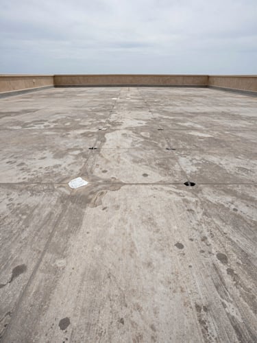 Wide environmental shot of a flat rooftop terrace under overcast Mediterranean sky, visible water staining and membrane blistering across the surface, drainage points clogged, parapet wall at the far edge, no people, flat diffused daylight showing surface condition