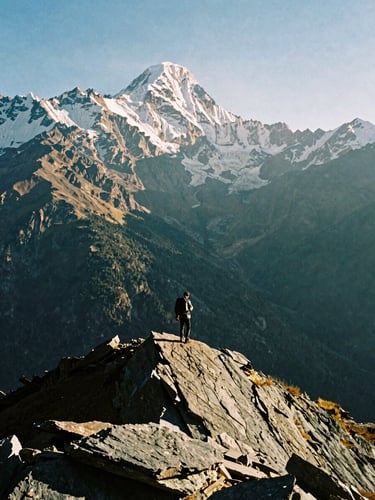 Dramatic high-contrast 35mm film shot of a lone trekker standing on a sharp slate ridge in the Indian Himalayas, golden-hour low-angle sunlight casting long shadows, massive snow-capped peaks towering in the background.