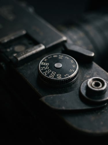 Extreme close-up of a vintage rangefinder camera top plate, focus on the shutter speed dial, low-key side lighting on deep charcoal metal, shallow depth of field