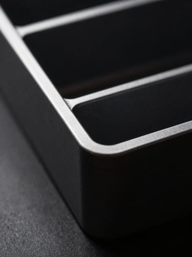 Macro detail of a dark anodized aluminum desk organizer, sharp highlights, dramatic side-lighting, deep shadows