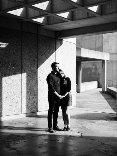 High-contrast black and white photograph of a modern couple standing in a minimalist concrete pavilion, sharp geometric shadows, dramatic side lighting, 35mm film grain.
