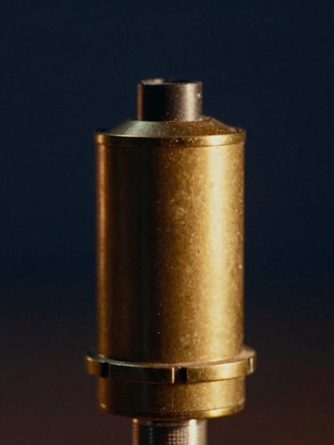 Tight geometric close-up of a vintage brass microphone capsule, warm golden-hour studio lighting, soft shadows, deep navy background, 35mm film grain.