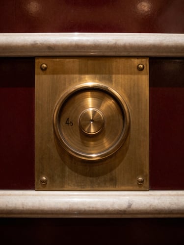 Extreme close-up of a polished brass elevator dial and marble trim in a five-star hotel lobby, warm interior glow, sharp architectural shadows, 35mm.