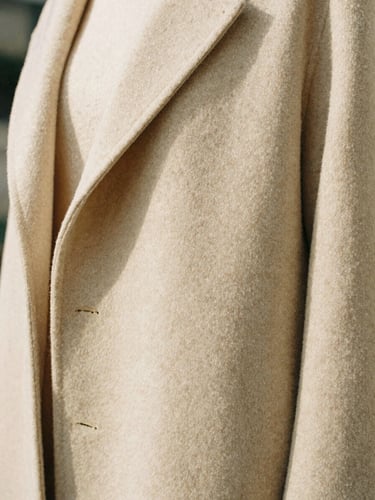 A close-up of a structured cream wool coat drape, architectural folds in soft natural daylight, shallow depth of field, warm sand tones.