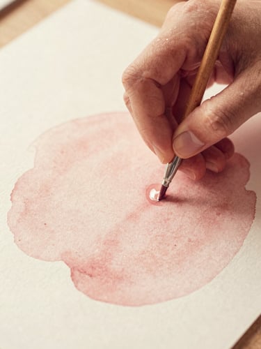 Close-up of artist's hands mixing a soft dusty rose wash with water, delicate paper grain visible, warm diffused golden-hour light
