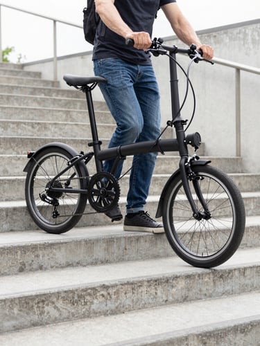 Medium-close shot of a commuter carrying a neatly folded black e-bike up concrete subway stairs, bright natural overcast daylight, soft shadows, clean urban background.