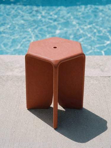 Low-angle architectural shot of a terracotta red monobloc stool casting a sharp shadow on a raw concrete pool deck under harsh midday sun, sparkling blue water in the background, crisp architectural photography.