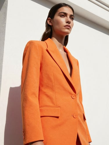 Low-angle heroic shot of a model in a structured vivid orange blazer against a crisp white architectural wall, direct bright sunlight, sharp high-contrast shadows