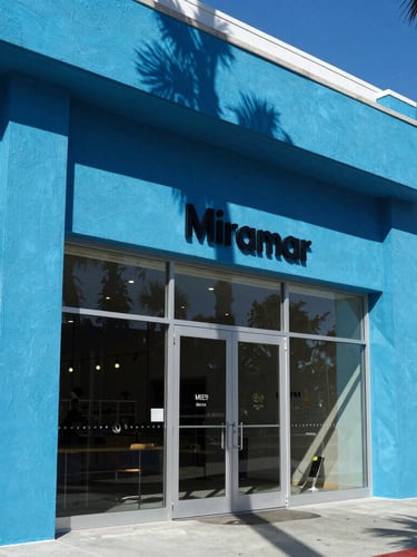 Low-angle heroic shot of a modern architectural storefront in Miramar Beach, bright direct sunlight, sharp electric blue shadows.
