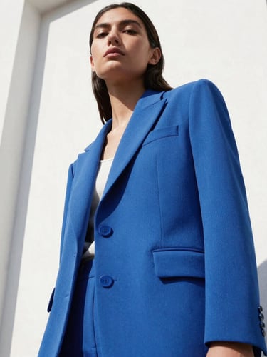 Low-angle heroic shot of a model in a structured electric blue blazer against a crisp white architectural wall, direct high-contrast sunlight with sharp shadows