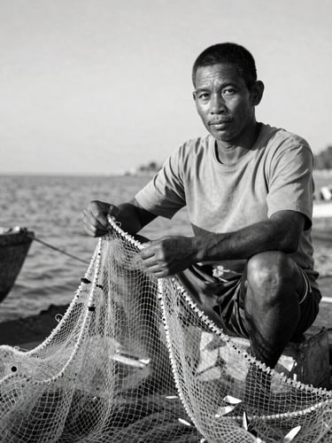 Black and white environmental portrait of a fisherman mending nets, natural daylight, deep shadows, looking directly into the lens with quiet resolve.