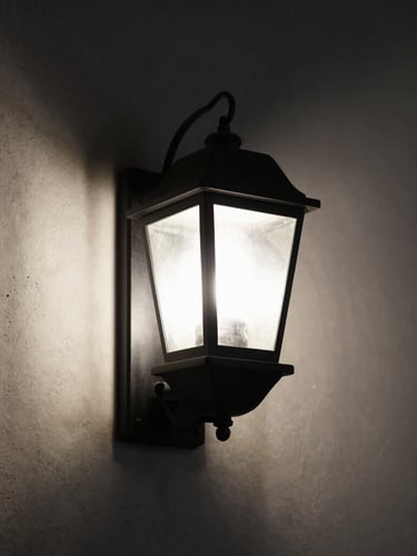 Moody close-up of a matte black iron wall lantern casting a precise, low-glare pool of light against a raw plaster wall, directional side-lighting, deep shadows, 50mm lens