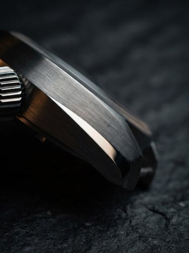 Extreme macro close-up of a brushed steel watch case, directional side-lighting catching the polished bevels, dark slate background, moody high-contrast details