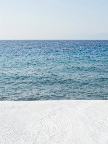 Minimalist coastal horizon of the Aegean sea, soft water movement, clean bright daylight, whitewashed plaster texture in the foreground, serene and restorative atmosphere.