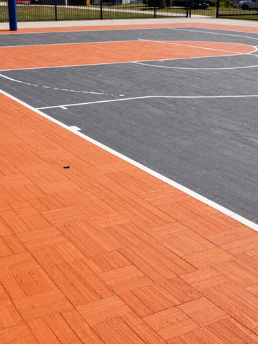 Sharp architectural sports photography of a freshly completed outdoor basketball court with orange and charcoal PP interlocking tiles, bright crisp daylight, wide-angle lens